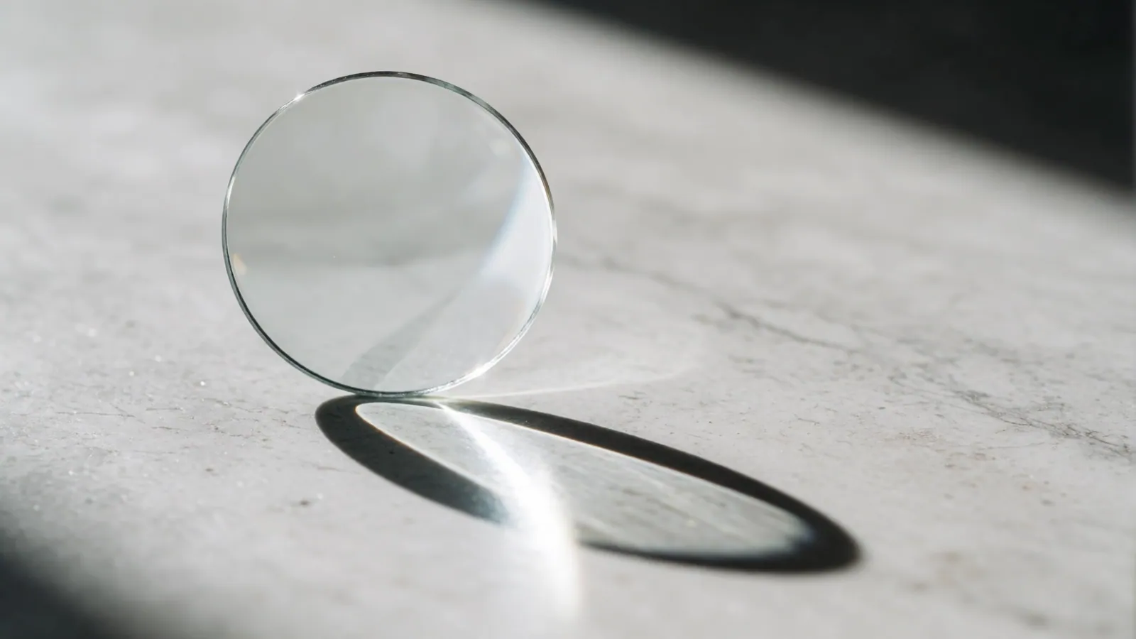 A single bare lens standing on pale stone, catching a crescent of morning light and casting a hard bright shadow.