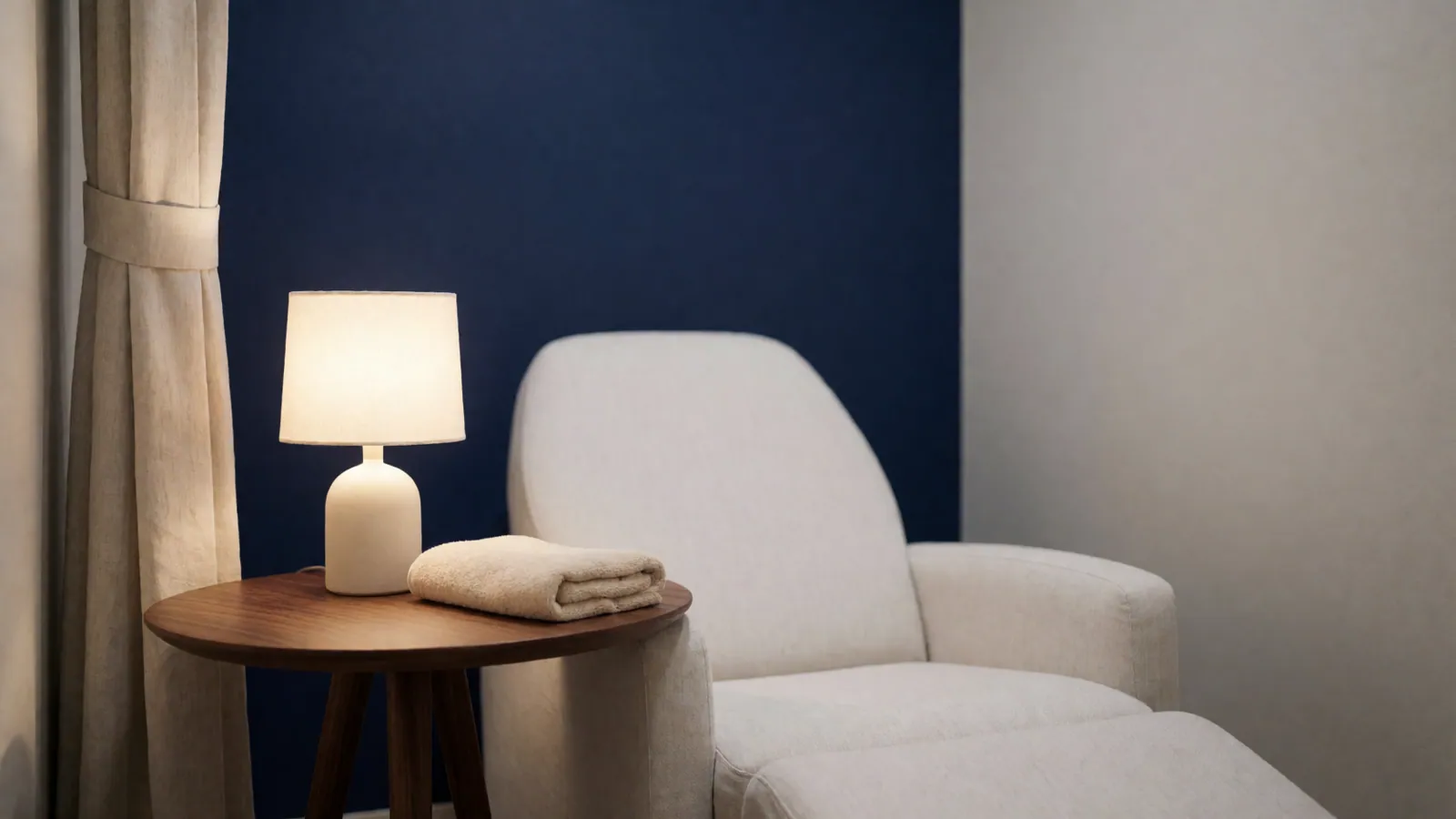 A treatment alcove: a pale linen chair, a side table with a folded towel and a lit lamp, a deep indigo wall and a half-drawn curtain