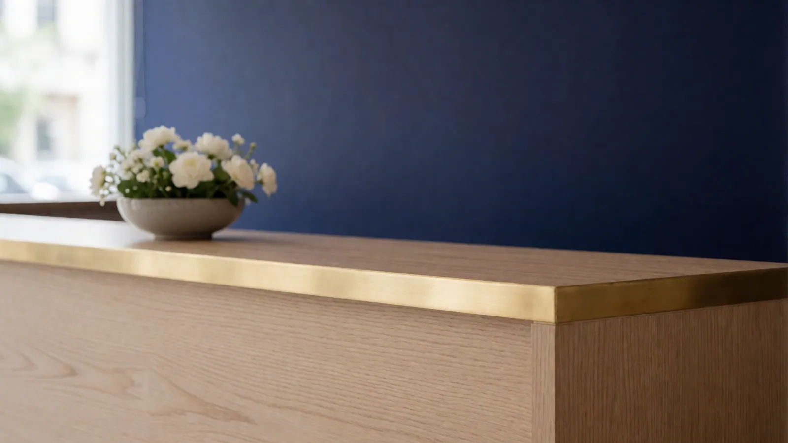 The reception counter in pale oak with a brass edge, a low bowl of white flowers, a deep indigo wall behind