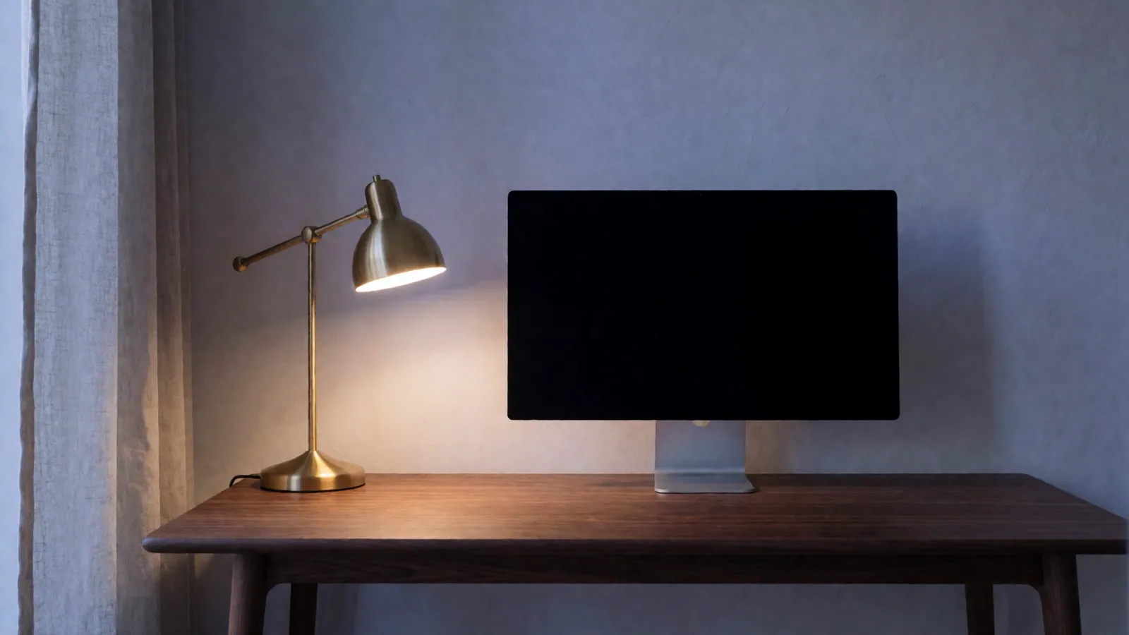 A home office corner at dusk: an empty walnut desk, a monitor switched off, a lit brass task lamp
